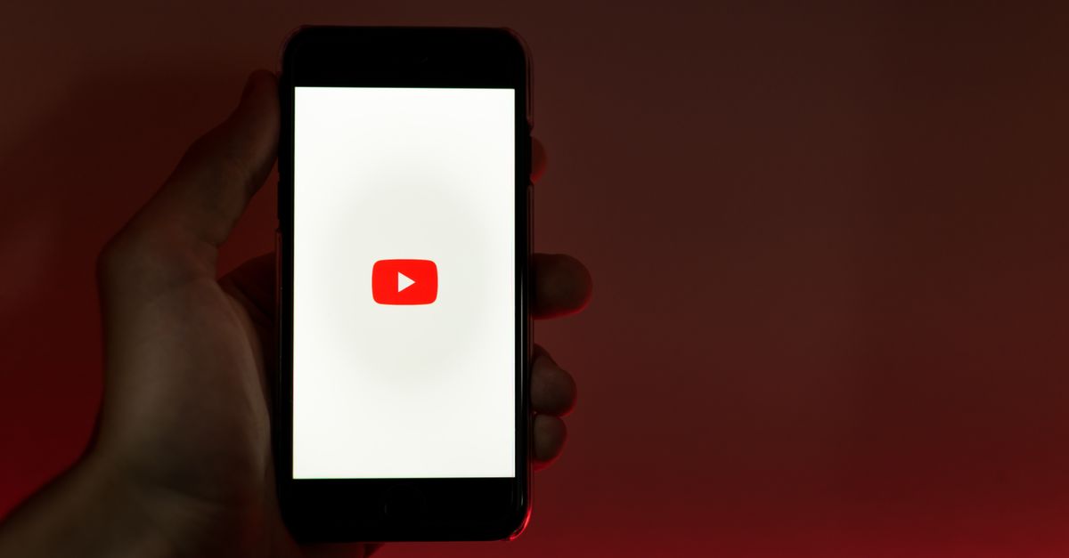 monetizar con videos en YouTube: A hand holding a smartphone displaying the YouTube app against a red background.