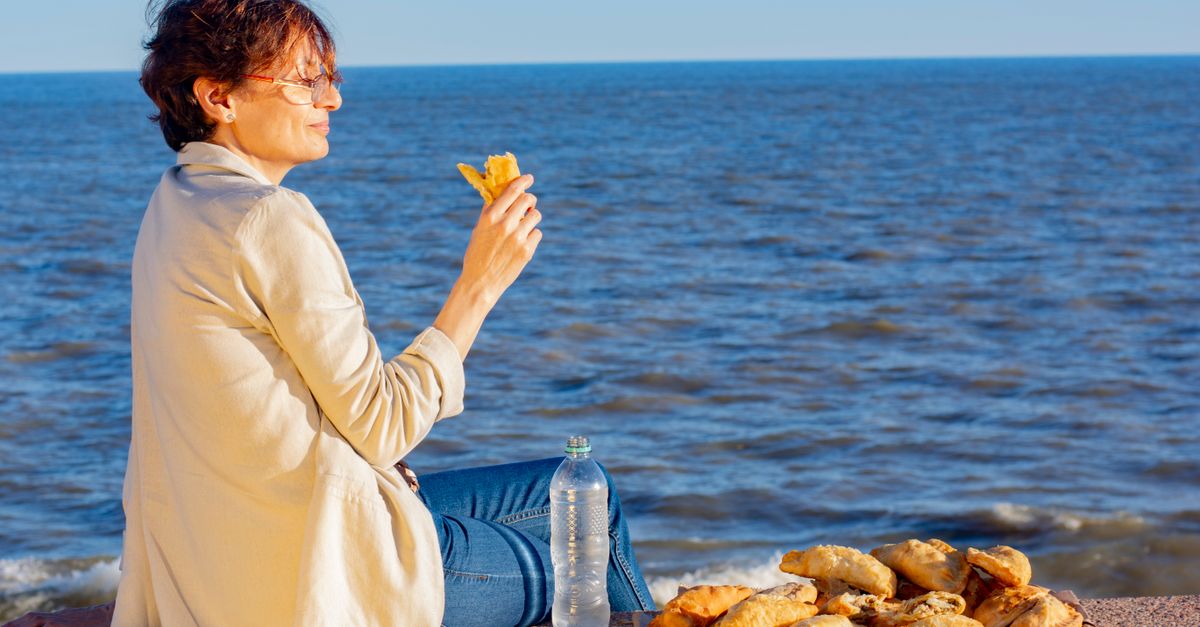 cómo prepararse para olas de calor sin descuidar la salud diaria: A woman enjoys empanadas by the ocean in Montevideo, Uruguay during a sunny day.