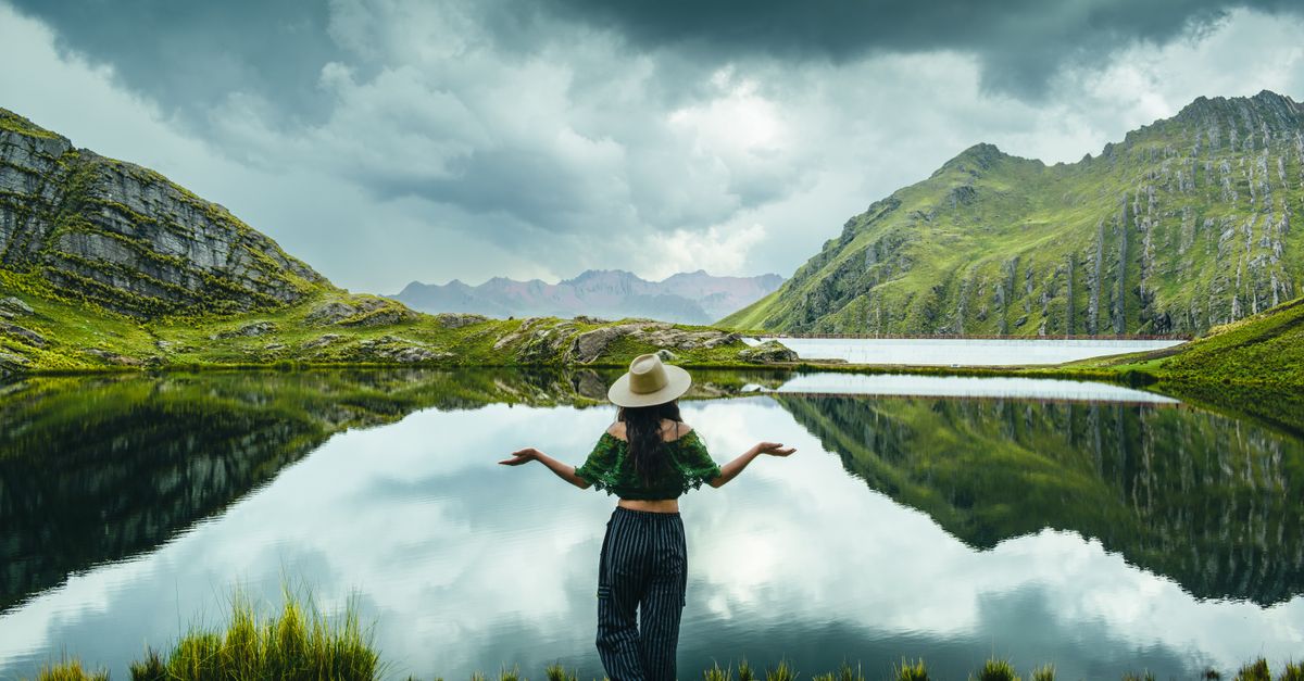 Vacaciones a destinos frescos: cómo planear un viaje cómodo, sostenible y fuera de temporada alta viajes inteligentes destination specific travel: Woman stands by a serene lake in Marangani, Cuzco, Peru, surrounded by lush green hills.
