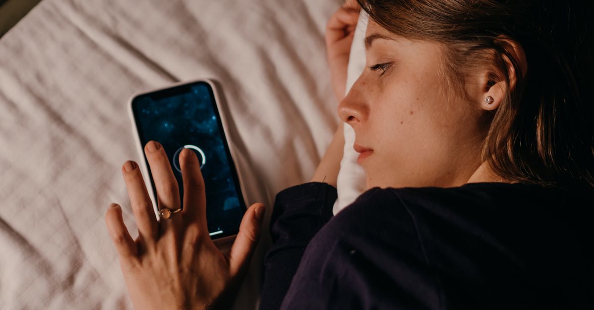 Cómo usar relojes y anillos inteligentes para dormir mejor sin obsesionarte con los datos cultura visual creatividad arte contemporáneo: A woman lying in bed, engaged with her smartphone, creating a tranquil nighttime scene.