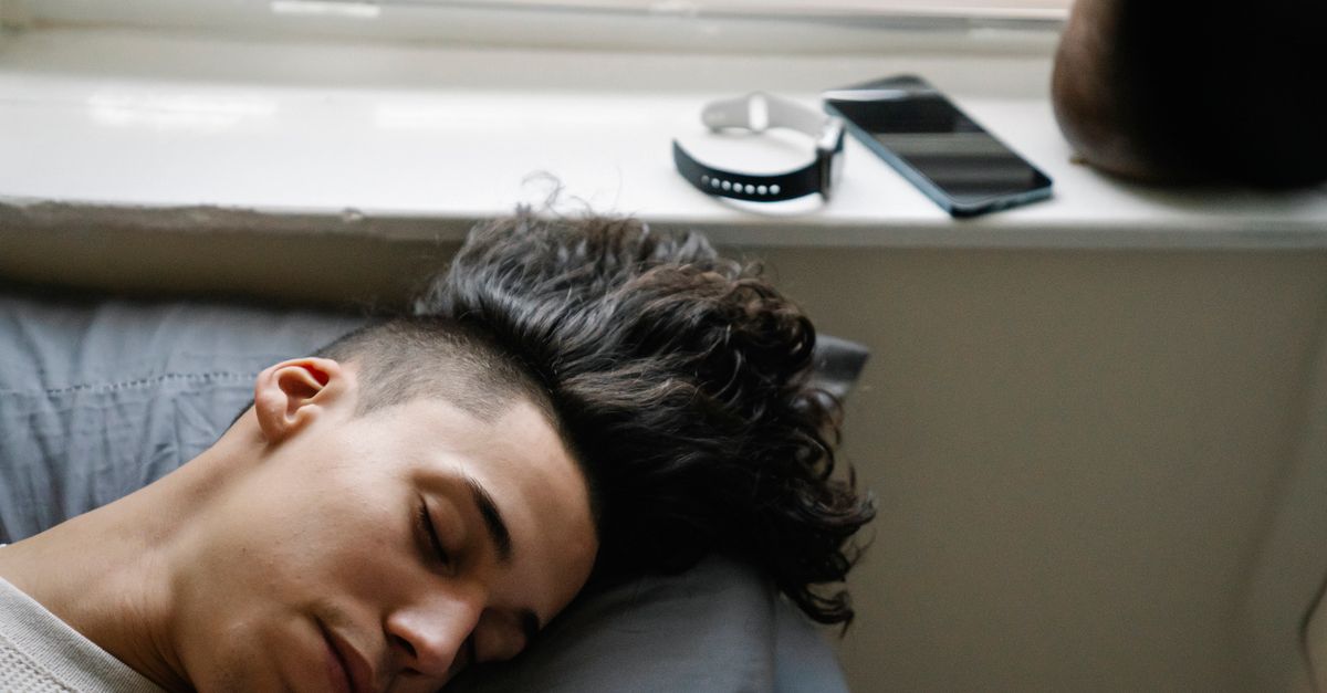Cómo usar relojes y anillos inteligentes para dormir mejor sin obsesionarte con los datos cultura visual creatividad arte contemporáneo: High angle of young Hispanic male with curly hair lying on bed near windowsill and sleeping in morning at home