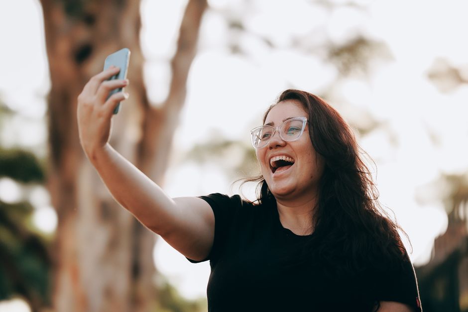 Smiling woman taking a selfie outdoors in a sunny park, capturing a joyful moment.